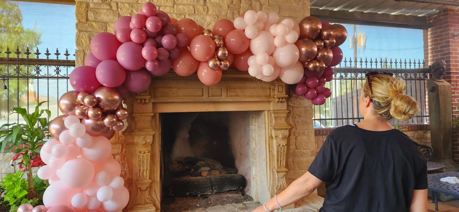 Organic blush, dusty mauve, and rose-gold chrome balloon garland with white daisies woven through, draped across the carved stone fireplace mantel and cascading down the left column on the patio at Farina's Winery & Cafe in Grapevine, TX.