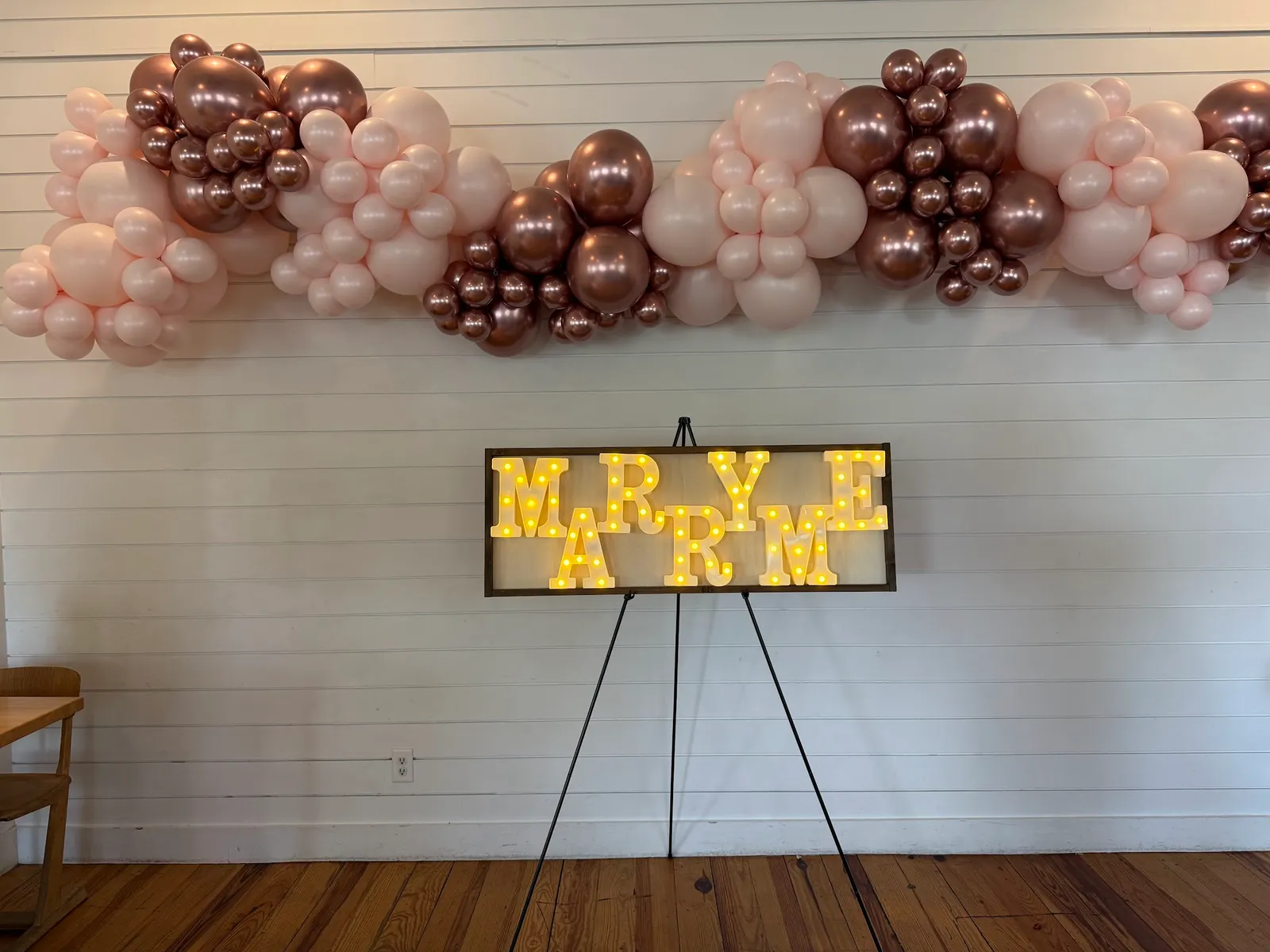 An organic balloon garland in rose-gold chrome, blush pink, and deep espresso-brown latex running along a white shiplap wall, with a wood-framed light-up marquee sign spelling MARRY ME on a black easel beneath it. An engagement celebration built inside George Coffee + Provisions in Old Town Coppell, TX.