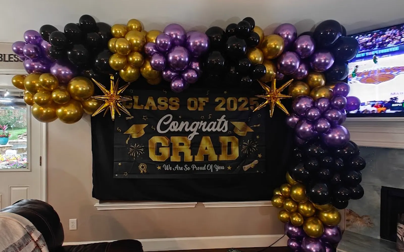 An organic balloon garland in chrome purple, gold, and black with gold foil starbursts framing a black Class of 2025 Congrats Grad backdrop, built for a Lamar Jackson themed high school graduation party in Corinth, TX.