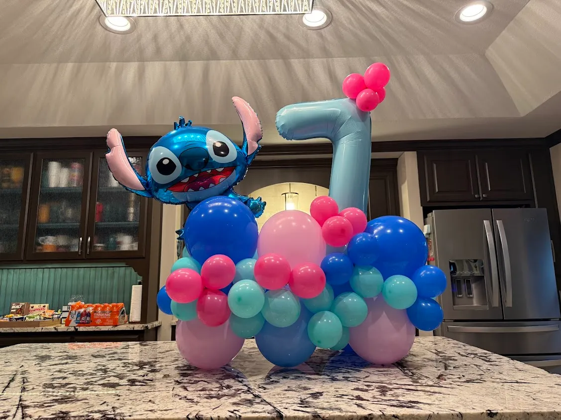A tabletop balloon centerpiece with a custom-ordered foil Stitch character and a light-blue number 7, on an organic cluster of royal blue, light pink, hot pink, aqua, and lavender balloons, built for a Lilo and Stitch 7th birthday in Coppell, TX.