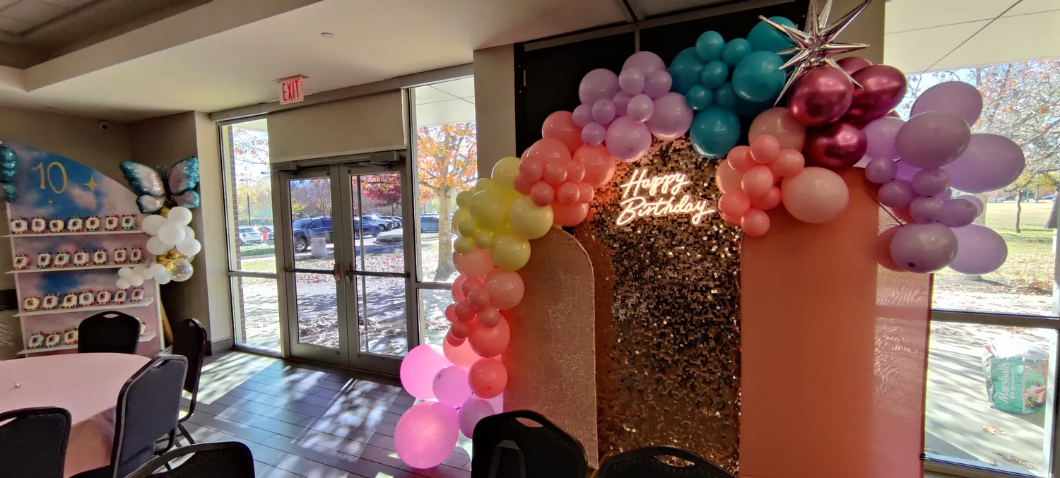 An organic balloon garland in mixed pink, coral, lavender, mint, butter yellow, chrome silver, and chrome rose-gold balloons of three different sizes, framing a rose-gold sequin Happy Birthday panel and a gold 10 foil number — an example of the organic balloon garland service from Pecan Creek Events.