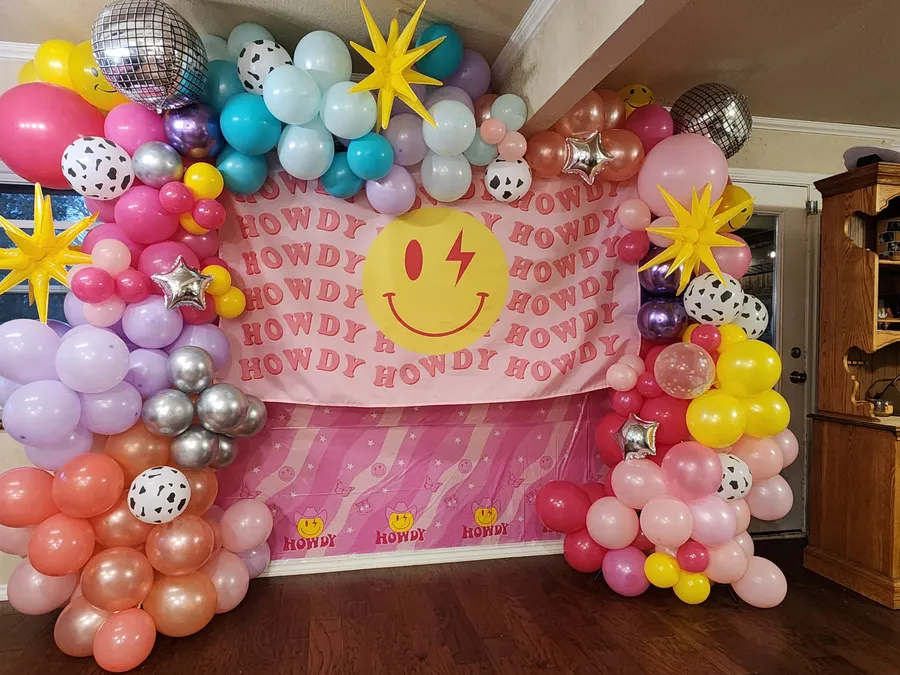 A pink HOWDY backdrop framed by a seven-color organic balloon arch with cow-print, chrome, and foil-starburst balloons and mirrored disco balls, set up for a Preppy Cowgirl 10th birthday at a home in Colleyville, TX.