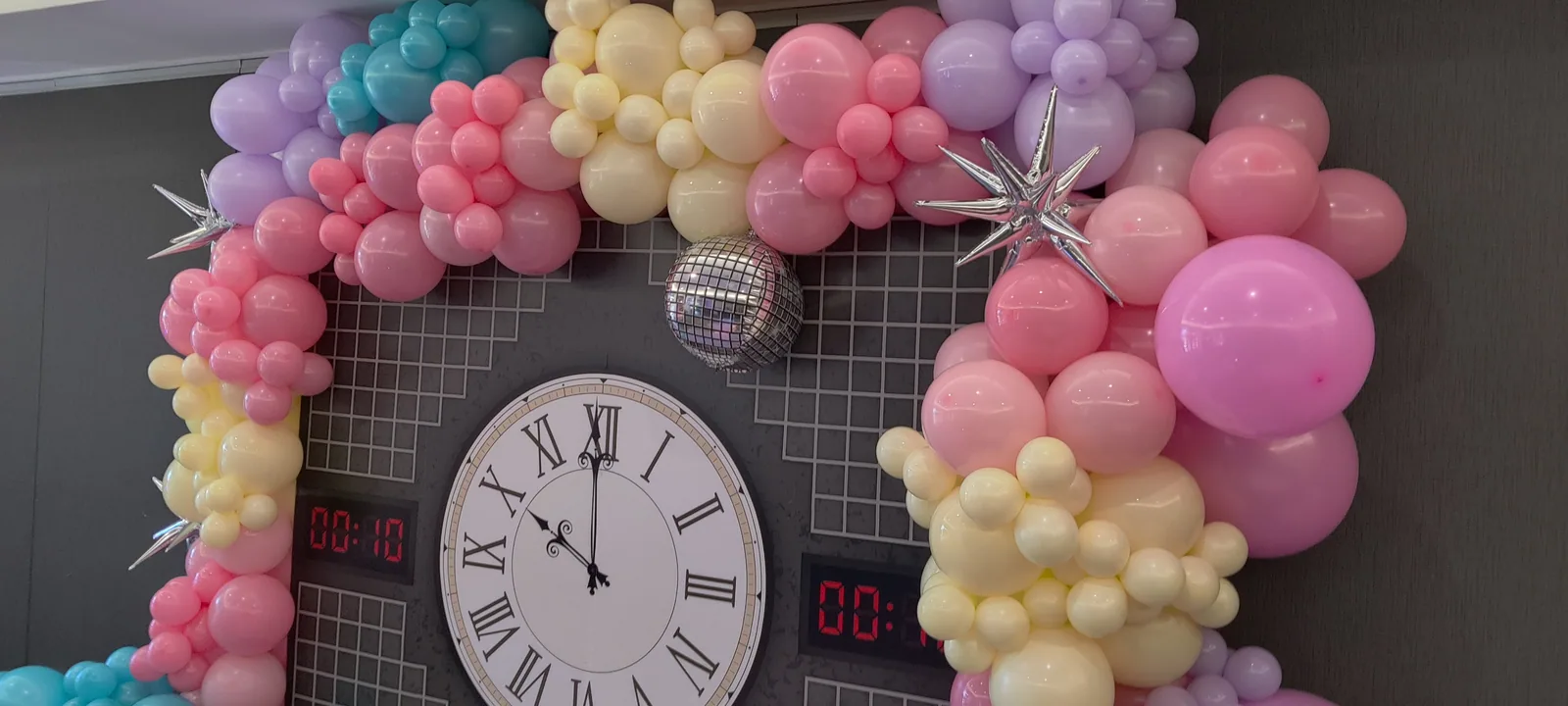 Pastel pink, lavender, mint, and yellow organic balloon garland framing a large analog clock with Roman numerals, digital LED displays reading 00:10 on each side, a silver disco ball, and silver starbursts — a Taylor Swift Midnights-themed birthday backdrop built at The Keller Pointe in Keller, TX.