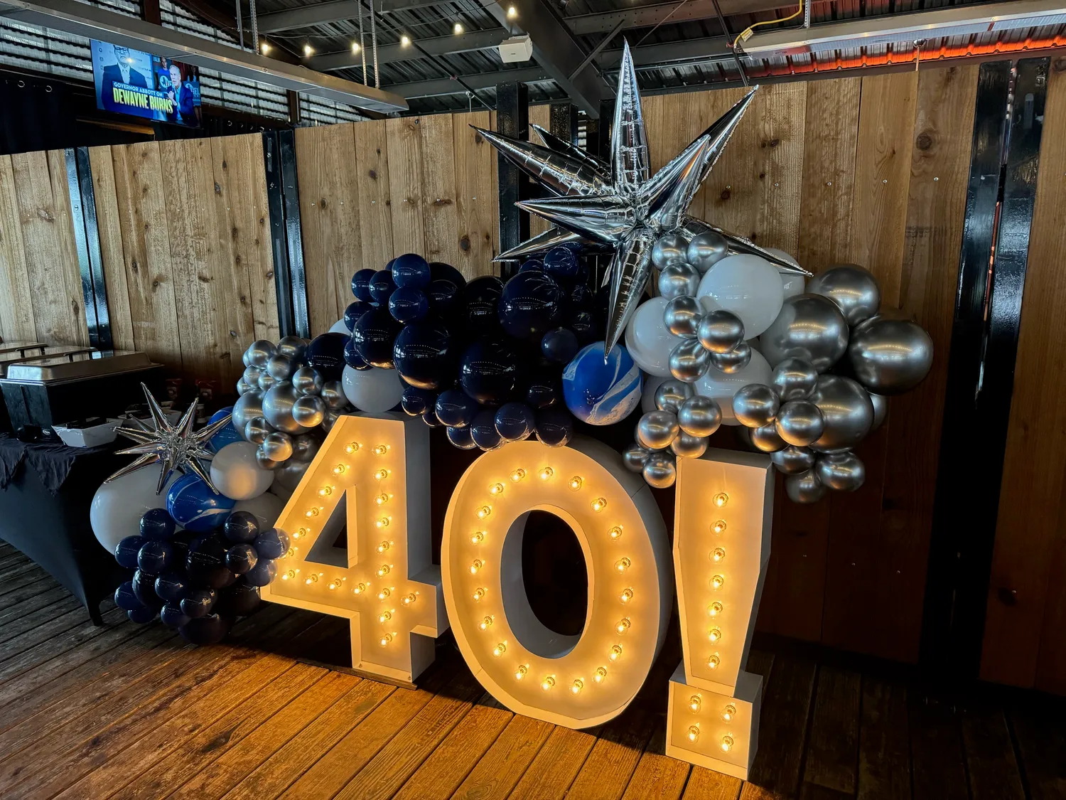 An illuminated three-foot 40 marquee letter installation as the anchor object of a themed event backdrop, surrounded by an organic balloon garland in navy, deep blue, and chrome silver with a silver foil starburst — a milestone birthday backdrop built by Pecan Creek Events.