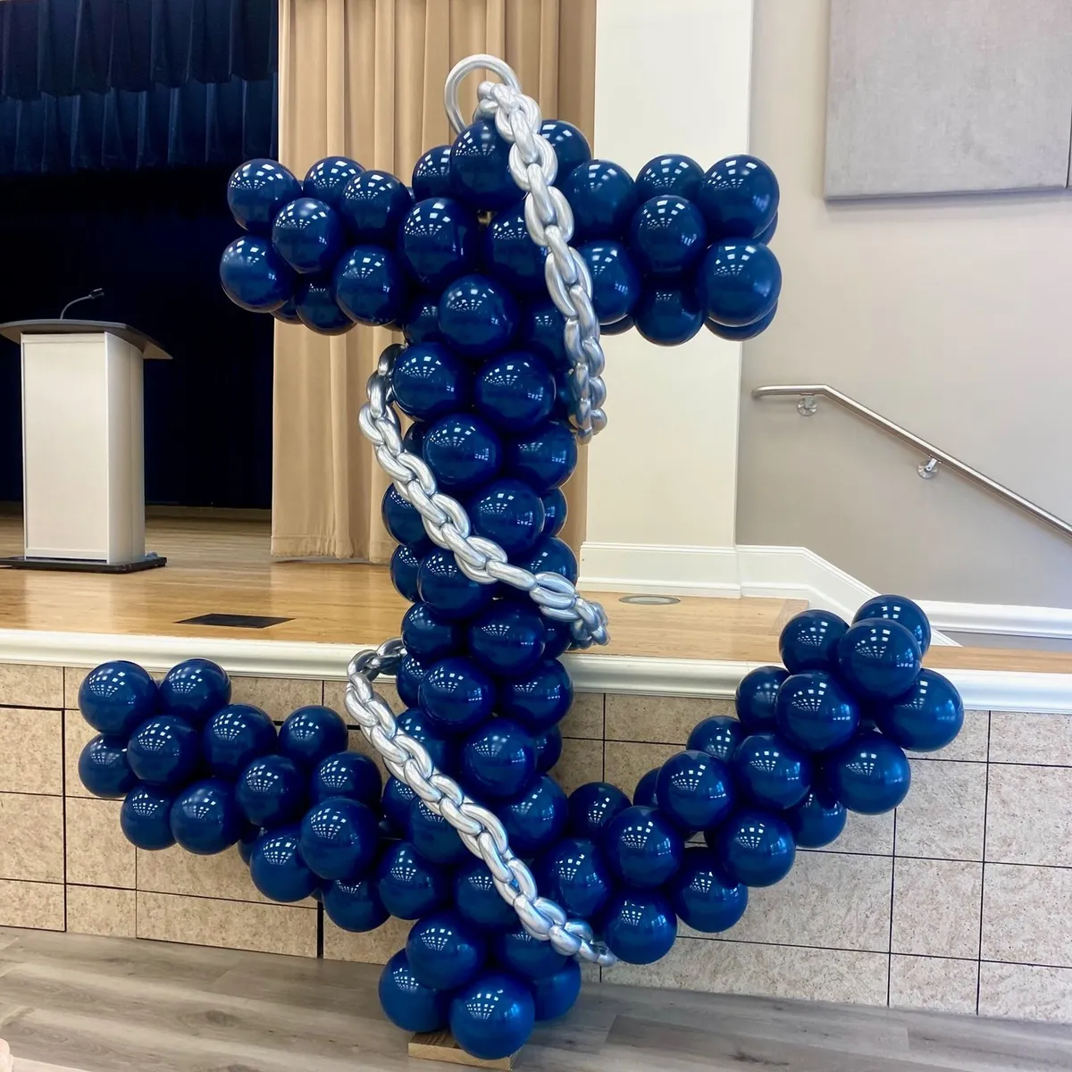 A freestanding balloon sculpture shaped like a ship's anchor in navy blue balloons with a wrapped silver balloon chain, built for Colleyville Elementary's Yacht Rock-themed PTA event in Colleyville, TX.