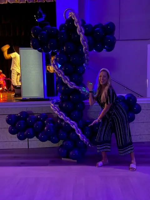 A guest posing beside the navy-and-silver anchor balloon sculpture under blue and purple event lighting at Colleyville Elementary's Yacht Rock PTA party.